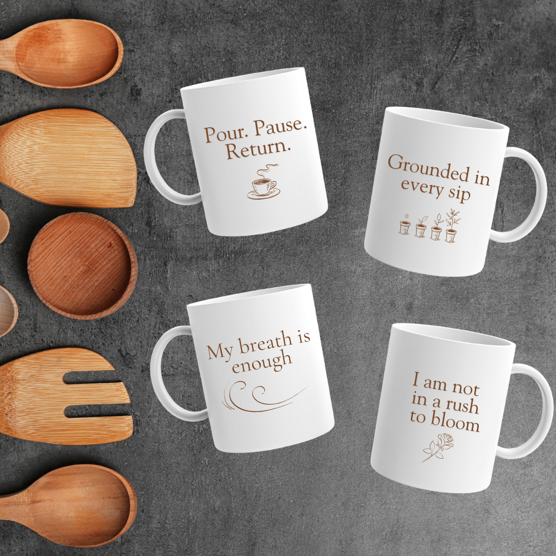 Four white mugs with brown affirmations and minimalist designs are arranged on a dark surface next to wooden utensils. Each mug features a calming phrase like “My breath is enough” and “Pour. Pause. Return.” in a soft, earthy aesthetic.